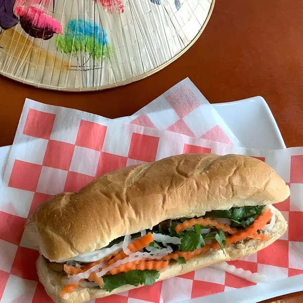 Zake's banh mi sandwich with carrots, daikon, and cilantro, on checkered paper, next to a conical hat.